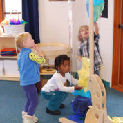 Children playing with a clear, pivoting Kodo Kids wind tunnel tube on a wooden base in a classroom setting.