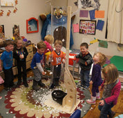 Clear pivoting wind tunnel with a light wood base, filled with floating packing peanuts, surrounded by excited children.