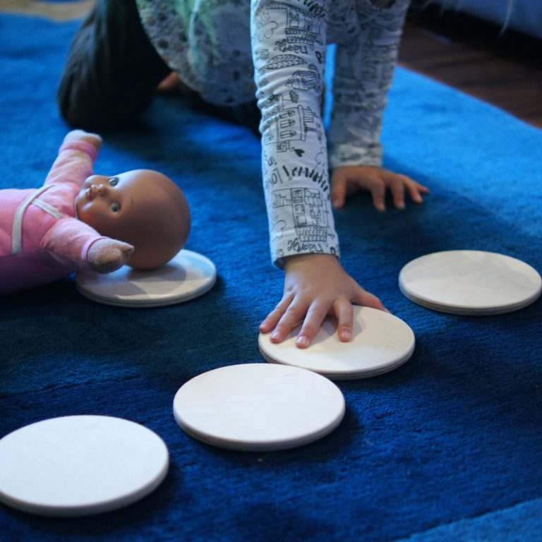 Child playing with smooth, circular natural wood biscuits arranged on a blue carpet alongside a doll.