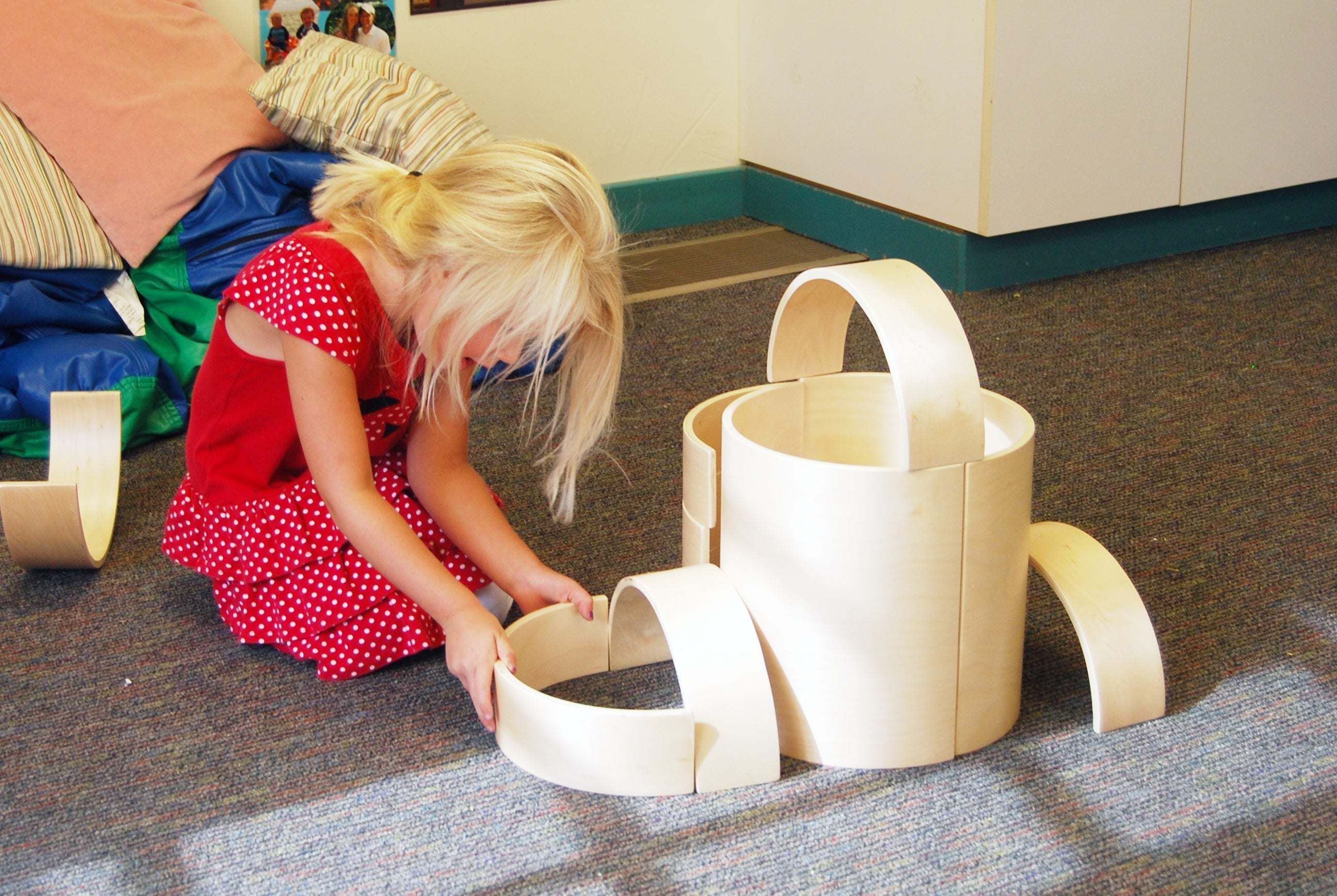 A young girl playing with a set of natural birch wood nesting arches on a carpeted floor.
