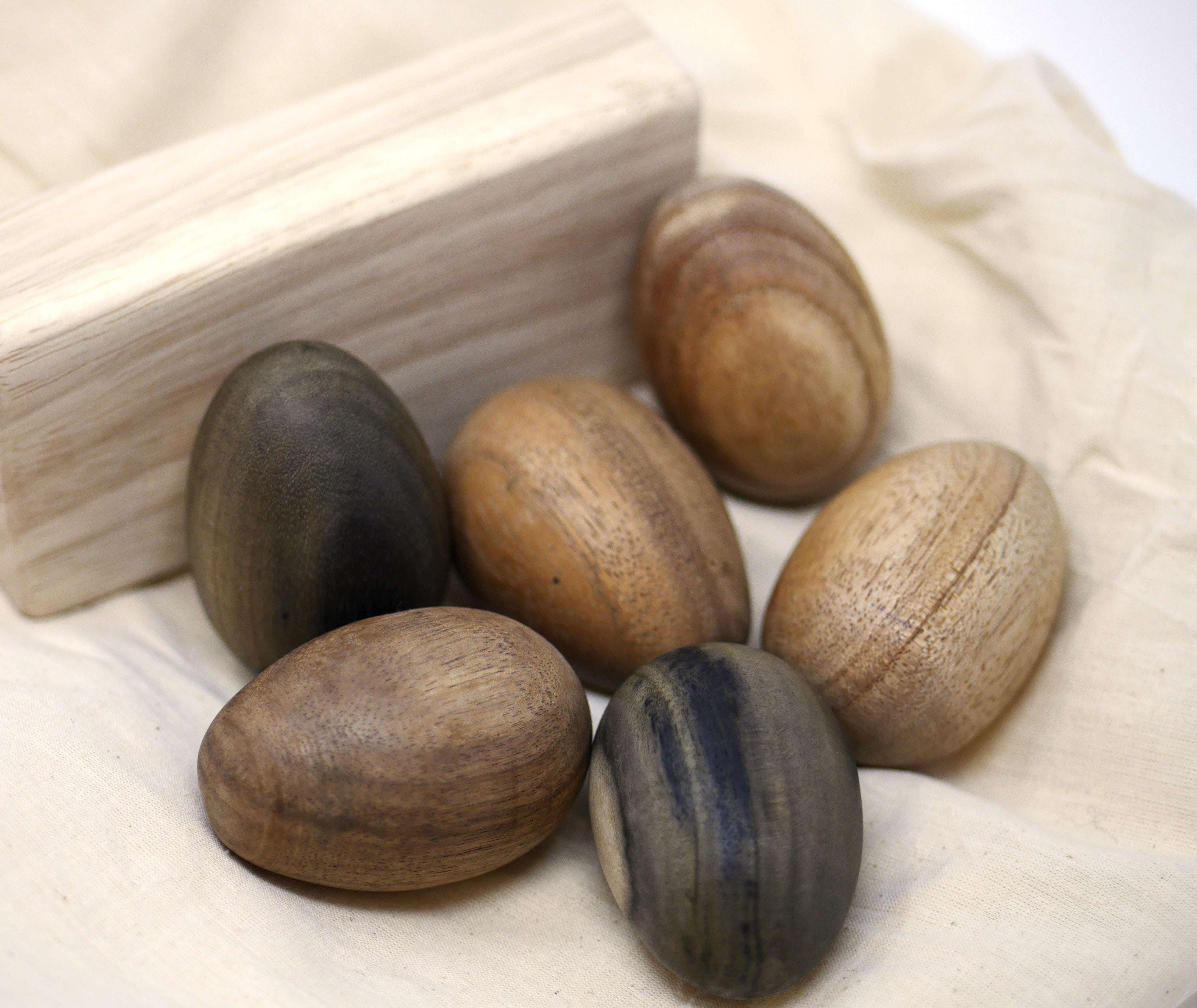 Set of six smooth, handcrafted wooden eggs with varied natural wood grains and tones resting on a cream fabric cloth.
