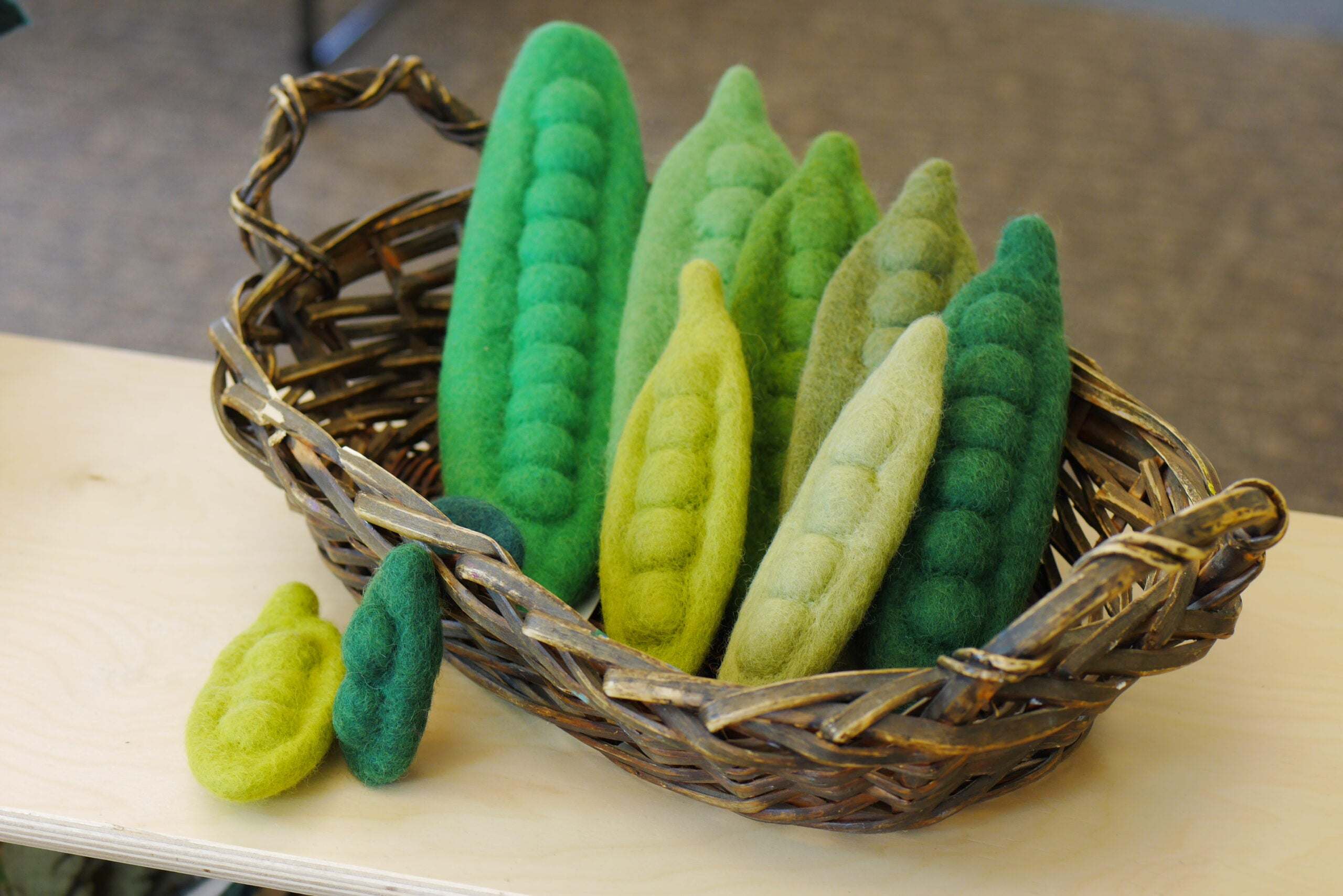 Ten handmade felted wool bean pods in various green shades arranged in a rustic woven basket.