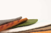 Stacked wool playscape mats in earthy tones of gray, orange, green, and white with a soft, textured felt finish.