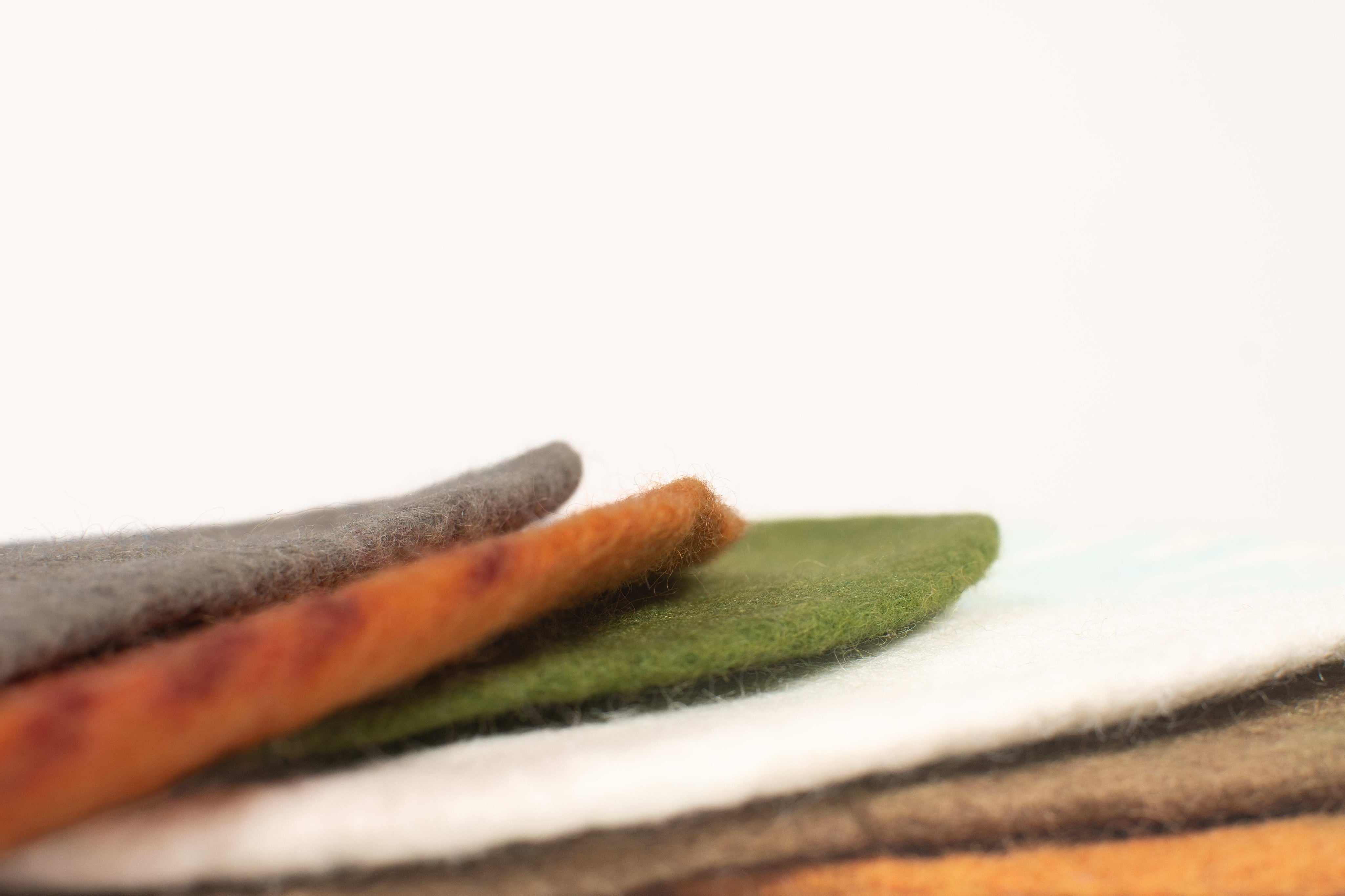 Stacked wool playscape mats in earthy tones of gray, orange, green, and white with a soft, textured felt finish.