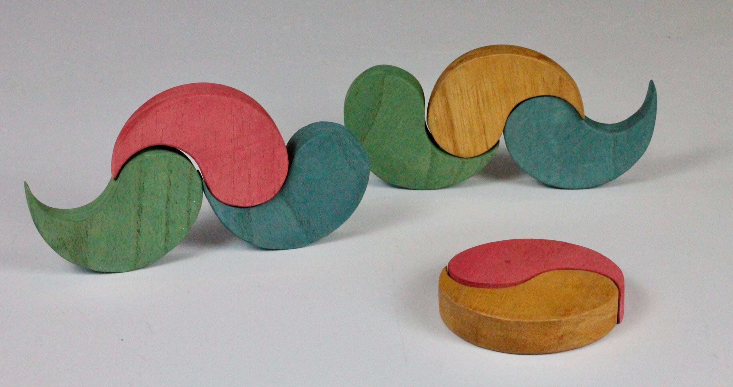 Handmade wooden Yim Yams puzzle pieces in earth tones, arranged in flowing shapes and a circular stack on a white background.